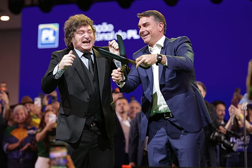 O senador brasileiro Flávio Bolsonaro, à direita, segura uma tesoura gigante, acompanhado pelo presidente argentino Javier Milei, durante a convenção do Partido Liberal 