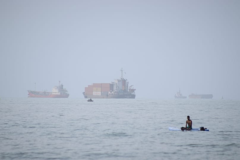 A man sits on a surfboard as a container ship and other commercial vessels appear anchored in the Strait of Hormuz off Bandar Abbas, Iran, Monday, July 27, 2026.