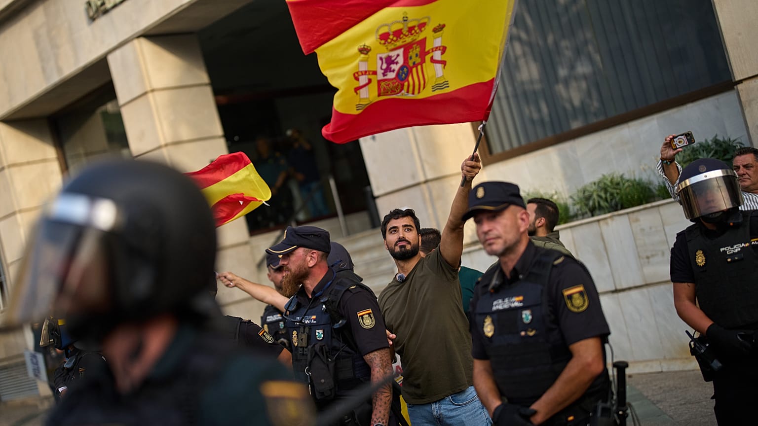El político de extrema derecha Alvise Pérez ondea una bandera española bajo protección policial mientras los vecinos de Ceuta protestan por su presencia, el sábado 1 de agosto