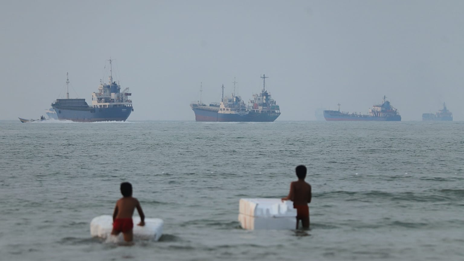 Two boys stand in shallow water with foam floats as cargo ships and other vessels are seen in the Strait of Hormuz off Bandar Abbas, 30 June, 2026