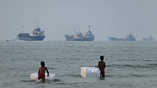 Two boys stand in shallow water with foam floats as cargo ships and other vessels are seen in the Strait of Hormuz off Bandar Abbas, 30 June, 2026