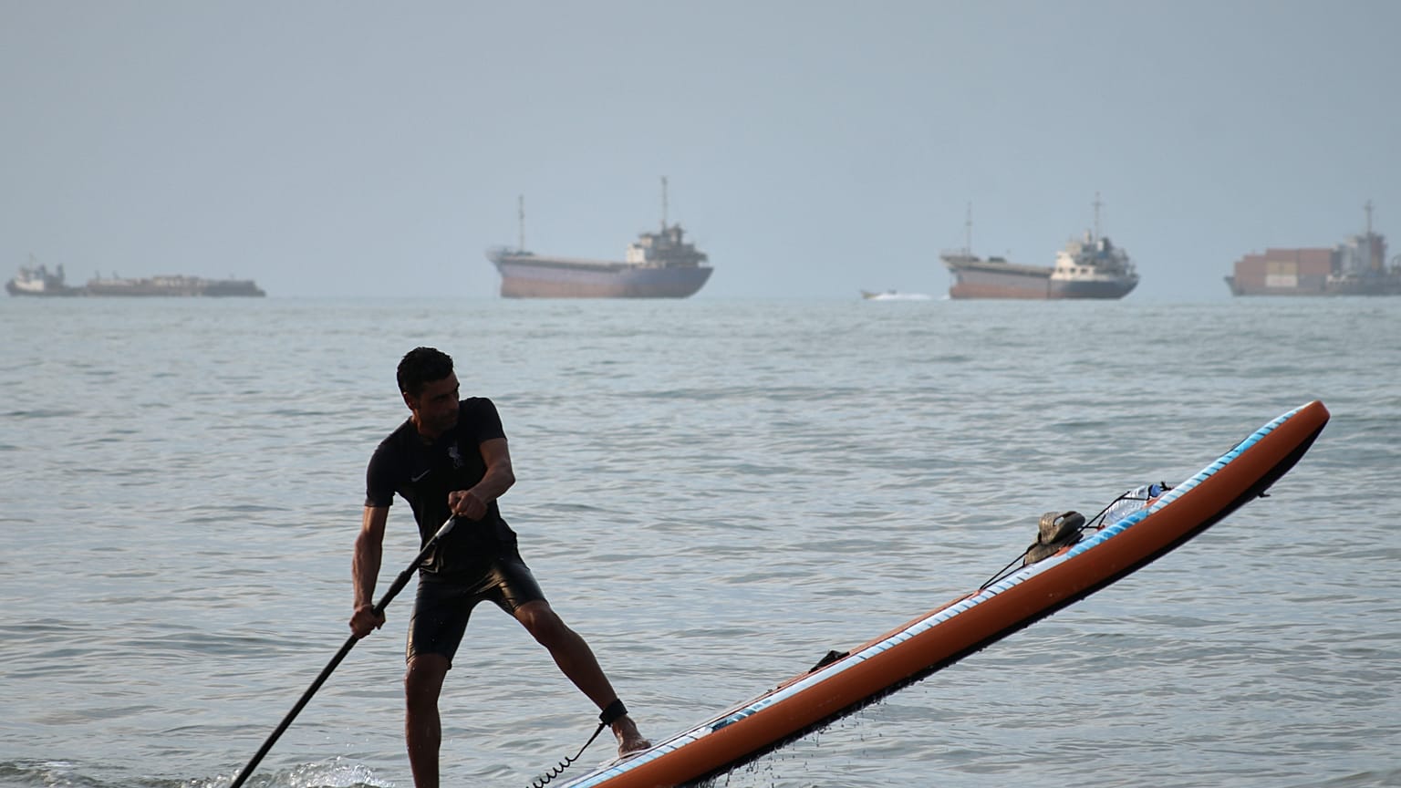 A man paddles a stand-up paddleboard as cargo ships and other commercial vessels appear anchored in the Strait of Hormuz off Bandar Abbas, Iran, Monday, July 27, 2026