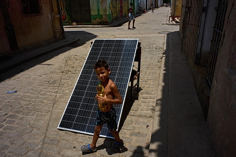A child walks with a bottle of oil past a solar panel set up on the street to charge batteries during a blackout in Havana, Cuba, Monday, July 6, 2026. 