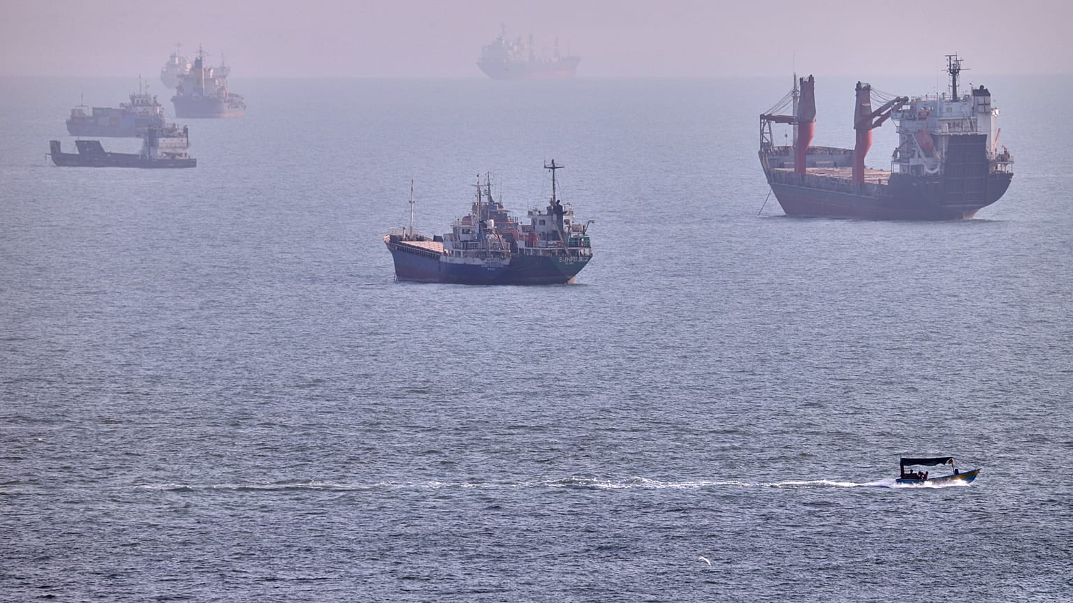 Ein kleines Boot fährt an Frachtern und anderen Handelsschiffen vorbei, die am fünften August 2026 vor Bandar Abbas in der Straße von Hormus ankern.
