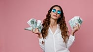 Une femme portant des lunettes de soleil tient des billets dans chaque main sur un fond rose.