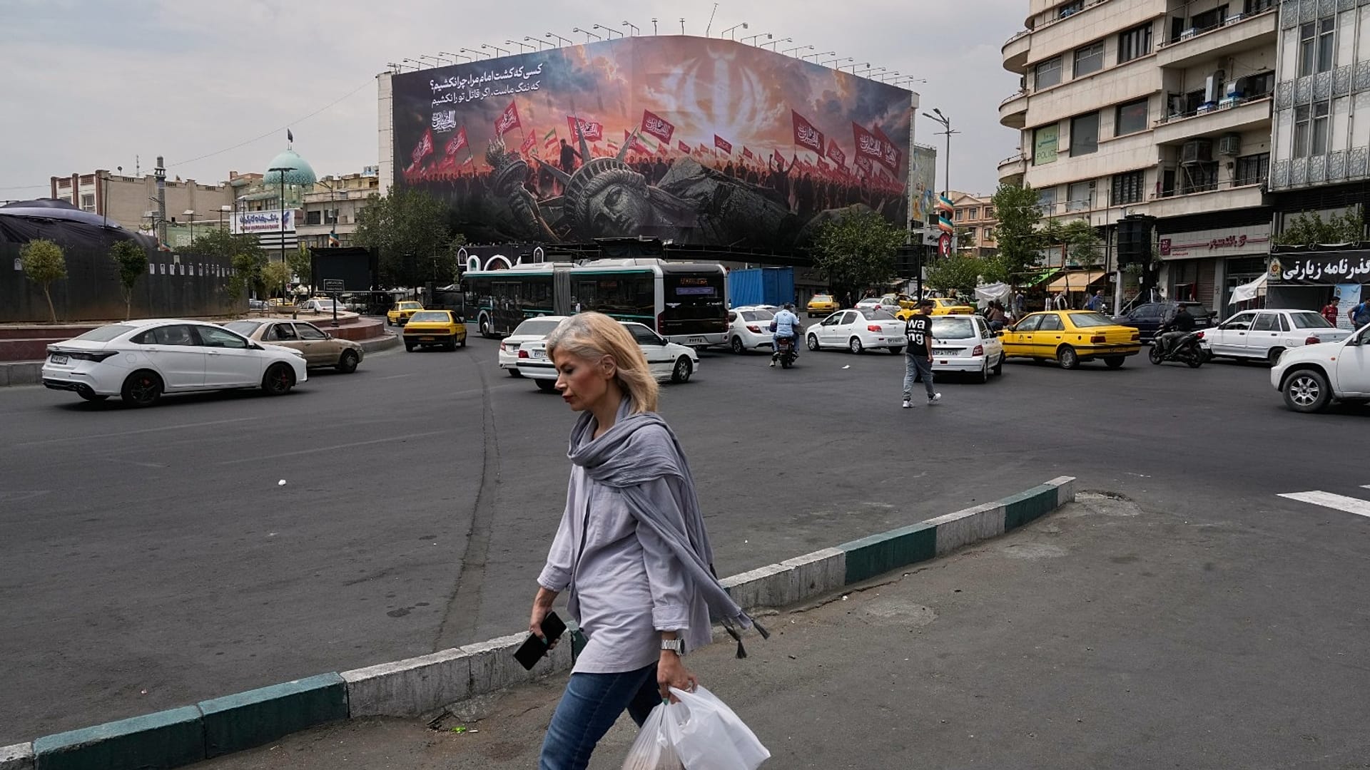 امرأة تسير في شارع بطهران، بينما تظهر خلفها لوحة لتمثال الحرية ساقطاً وحشود تحمل أعلاماً إيرانية، في ميدان الثورة.