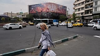امرأة تسير في شارع بطهران، بينما تظهر خلفها لوحة لتمثال الحرية ساقطاً وحشود تحمل أعلاماً إيرانية، في ميدان الثورة.