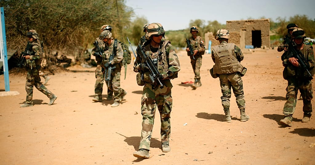 Soldiers in camouflage uniforms and helmets walking on a dirt road carrying rifles.