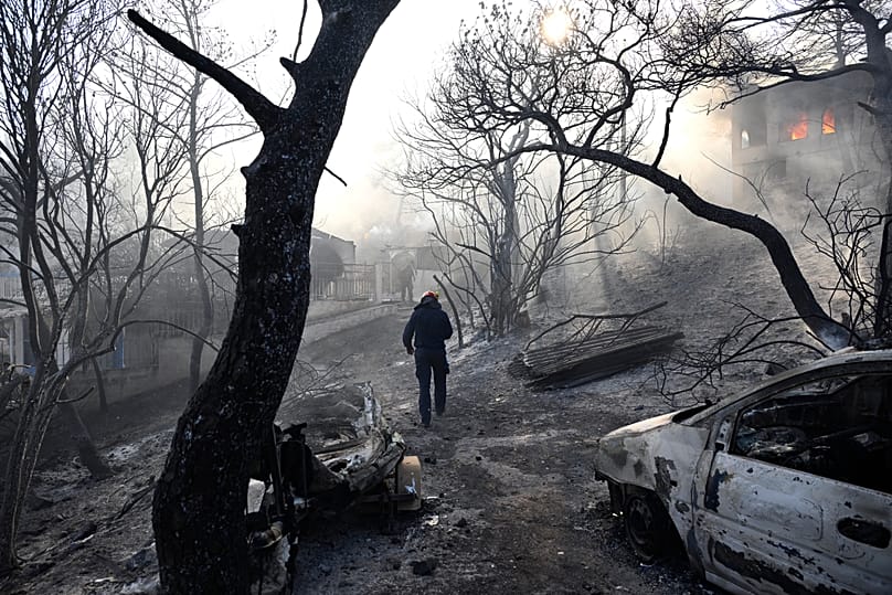 A firefighter walks through a burned area as flames engulf a house during a wildfire on the island of Salamina, 16 August, 2026