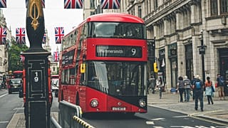 Iconic red double-decker bus on a busy street in London, England, with British flags overhead.