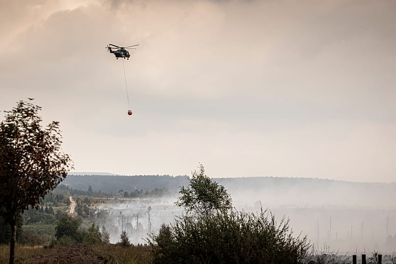 Ελικόπτερο ετοιμάζεται να αδειάσει κάδο Bambi Bucket με νερό πάνω από χωράφι, ενώ δασική πυρκαγιά καίει κοντά στο Waimes, 17 Αυγούστου 2026
