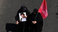 امرأة إيرانية تحمل ملصقًا للمرشد الأعلى آية الله مجتبى خامنئي بينما تحمل الأخرى علمًا يحتوي على كلمات تدعو المنتقمين للمرشد الأعلى الراحل آية الله علي خامنئي