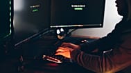 Силуэт человека за компьютером в тускло освещённой комнате, подчёркивающий угрозы кибербезопасности.