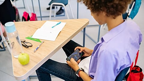 A child playing a mobile game at a school desk with school supplies nearby. 