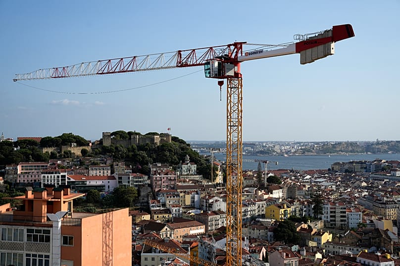 Ein Kran ragt über die Häuserdächer, im Hintergrund die Burg Castelo de São Jorge und der Fluss Tejo in Lissabon