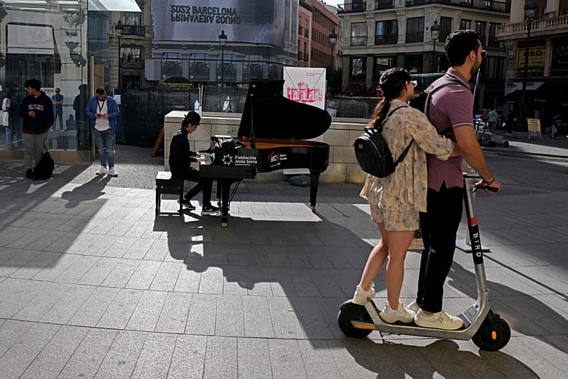 A man plays the piano in the street as a couple on an electric scooter ride by in downtown Madrid, 14 October, 2022