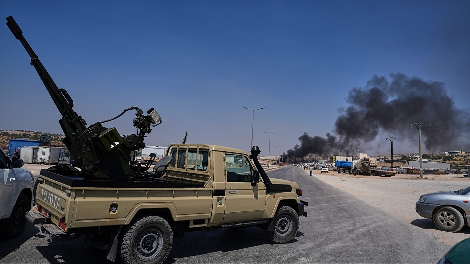 A pickup truck fitted with an anti-aircraft gun is parked across a road as smoke rises after an explosion at a weapons depot near Sarmada, 9 September 2026