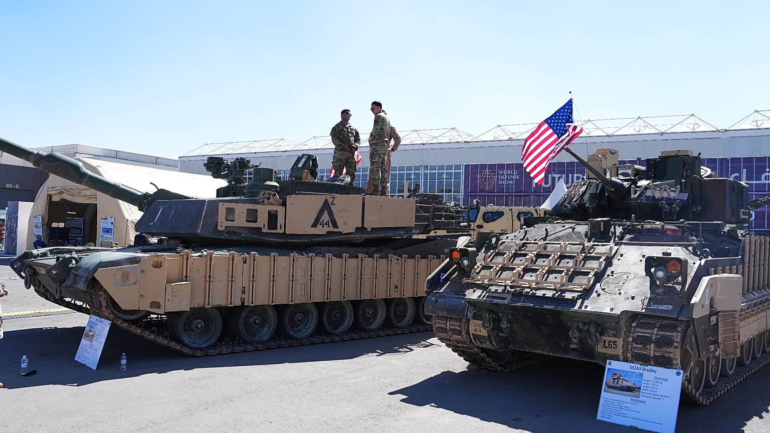 يقف أفراد من الجيش الأمريكي فوق دبابة القتال الرئيسية "إم 1 إيه 2 أبرامز" (M1A2 Abrams)، بينما تُعرض مركبة القتال المشاة "إم 2 إيه 3 برادلي" (M2A3 Bradley) في مكان قريب، وذلك 
