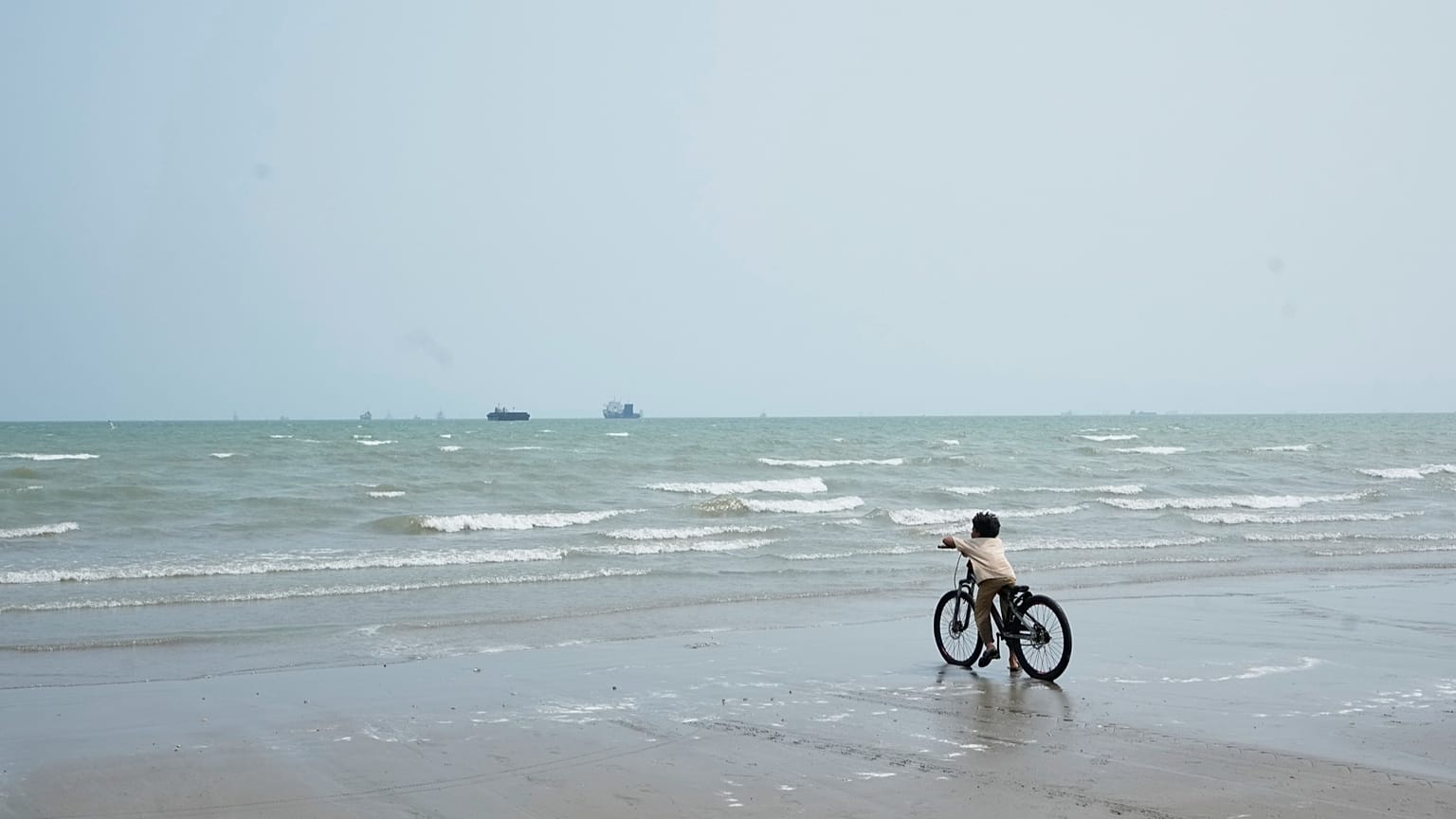 A boy pauses with his bicycle at the water's edge on a beach, looking toward commercial vessels anchored in the Strait of Hormuz off Bandar Abbas, 7 September, 2026
