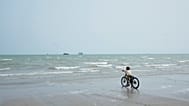 A boy pauses with his bicycle at the water's edge on a beach, looking toward commercial vessels anchored in the Strait of Hormuz off Bandar Abbas, 7 September, 2026