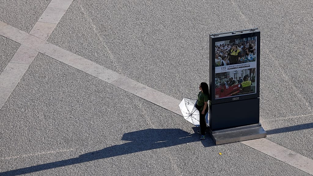 A person holds an umbrella, pausing in the shade of an information board while crossing Lisbon's Praça do Comércio on a hot September day.
