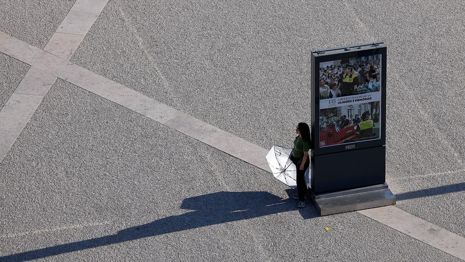 Eine Person hält einen Regenschirm und sucht den Schatten eines Infoboards. Sie überquert an einem heißen Septembertag den Praça do Comércio in Lissabon.
