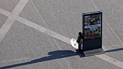 A person holds an umbrella, pausing in the shade of an information board while crossing Lisbon's Praça do Comércio on a hot September day.
