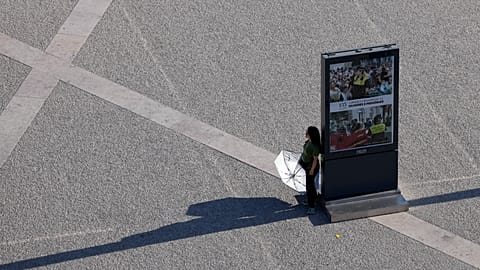 A person holds an umbrella, pausing in the shade of an information board while crossing Lisbon's Praça do Comércio on a hot September day.