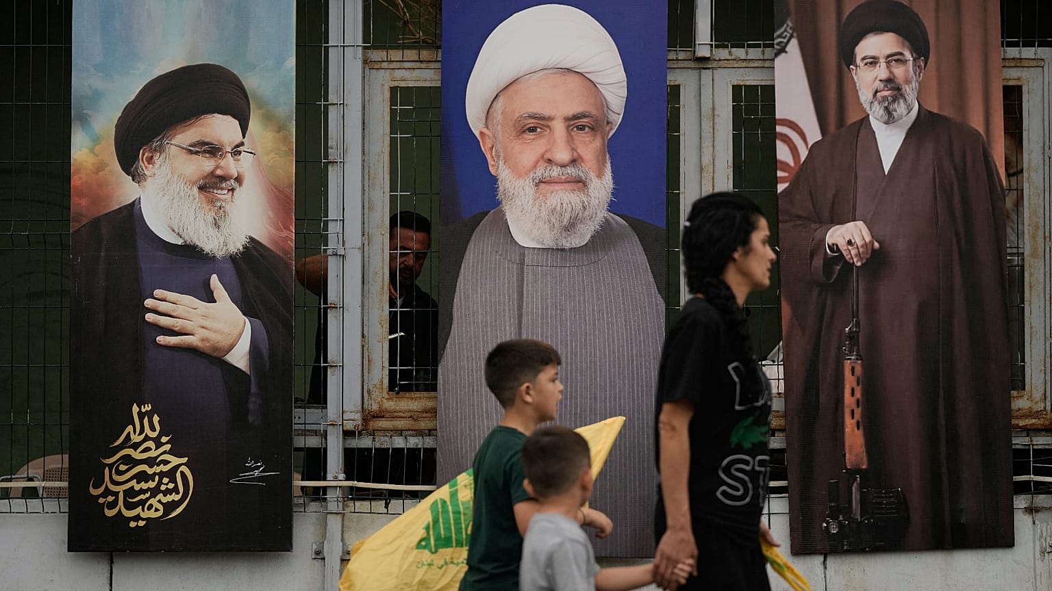 امرأة وأطفالها يحملون رايات "حزب الله" أثناء مرورهم أمام ملصقات تحمل صور كل من الأمين العام الراحل للحزب حسن نصر الله (يساراً)، والقيادي في الحزب نعيم قاسم (وسطاً)، والمرشد ال