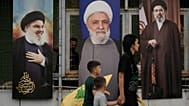 امرأة وأطفالها يحملون رايات "حزب الله" أثناء مرورهم أمام ملصقات تحمل صور كل من الأمين العام الراحل للحزب حسن نصر الله (يساراً)، والقيادي في الحزب نعيم قاسم (وسطاً)، والمرشد ال