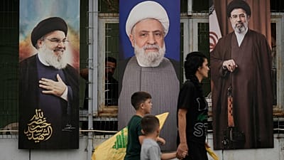 امرأة وأطفالها يحملون رايات "حزب الله" أثناء مرورهم أمام ملصقات تحمل صور كل من الأمين العام الراحل للحزب حسن نصر الله (يساراً)، والقيادي في الحزب نعيم قاسم (وسطاً)، والمرشد ال