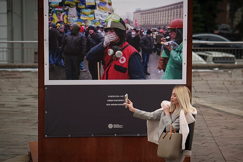 Женщина проходит мимо фотографии, на которой запечатлена сцена с Майдана 2014 года в Киеве, Украина, в четверг, 14 марта 2024 года, держа в руках мобильный телефон.