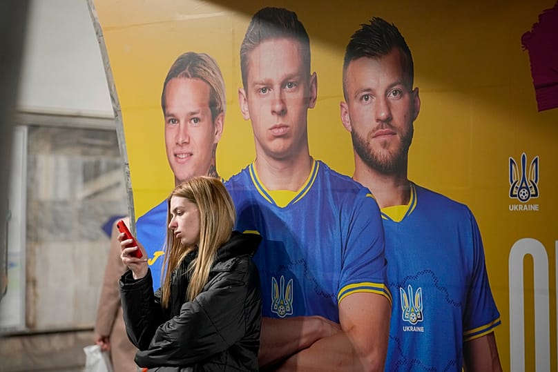 Молодая девушка проверяет свой мобильный телефон рядом с фотографиями украинских футболистов, представленными в рекламе на станции метро в Киеве, Украина, среда, 27 марта 2024