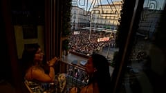 Dos mujeres se toman un refrigerio en uno de los comercios de la Puerta del Sol con la acampada de la vivienda como telón de fondo. 