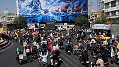 سائقو دراجات نارية أمام لوحة إعلانية تُظهر بطلاً أسطورياً فارسياً وشخصية طائرة تهاجم حاملات طائرات أمريكية، مصحوبة بعبارة "سيغرق جيش الشيطان في الخليج الفارسي" في طهران