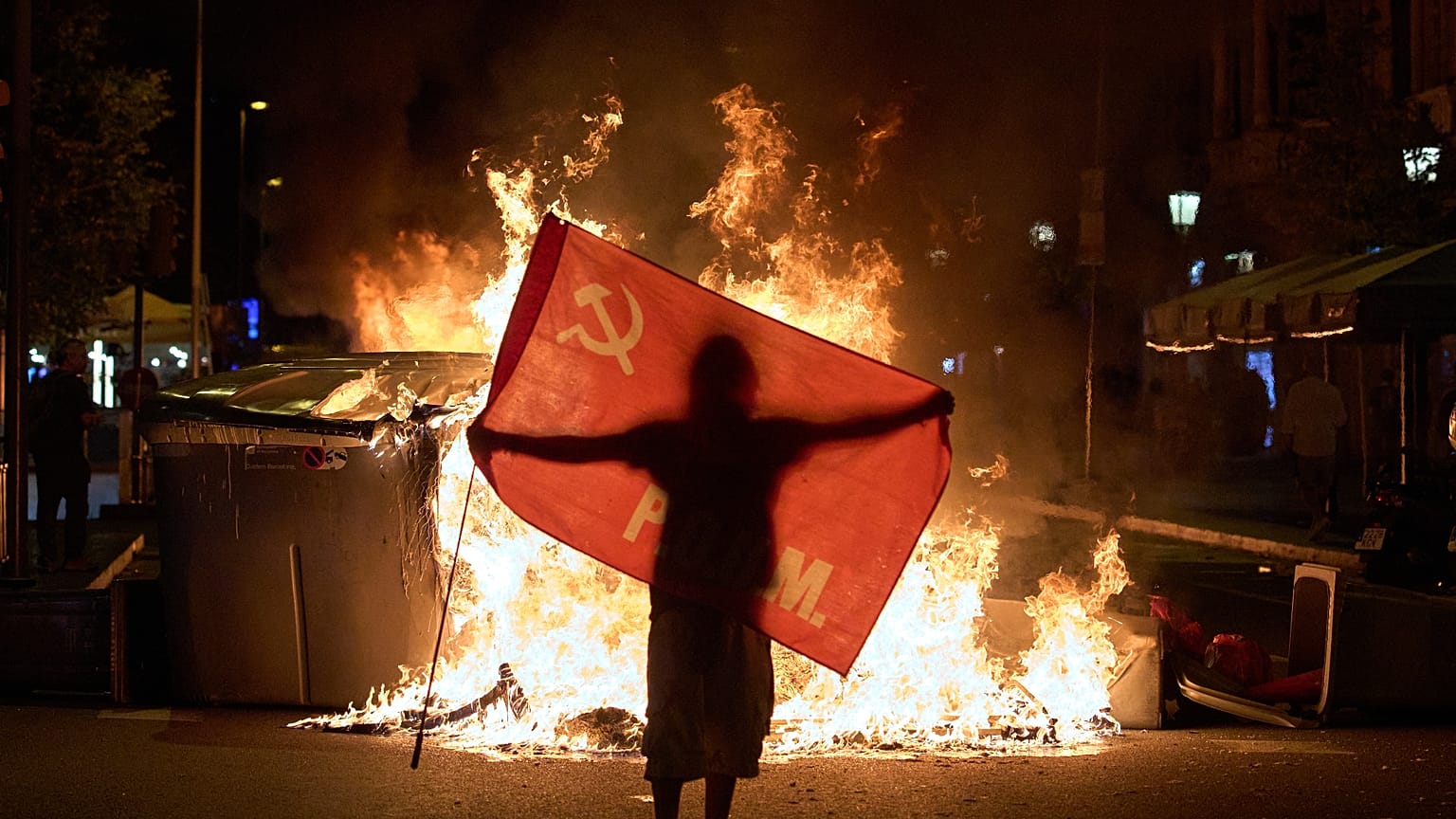 Ein Demonstrant hält eine Fahne neben brennenden Müllcontainern bei Zusammenstößen mit der Polizei in Barcelona, Spanien, am Montag, 5. Oktober 2026.