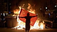 Протестующий держит флаг рядом с горящими мусорными баками во время столкновений с полицией после демонстрации, посвященной жилищному кризису в Испании. Барселона, 5.10.26г