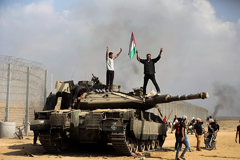 Palestinians celebrate by a destroyed Israeli tank at the southern Gaza Strip fence east of Khan Younis on Saturday, Oct. 7, 2023. (AP Photo/Yousef Masoud)
