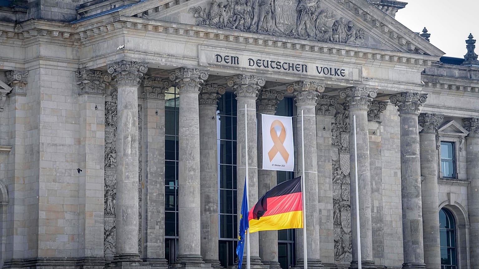 Ein Gedenkband mit der Aufschrift "7. Oktober 2023" hinter den auf Halbmast gehissten Flaggen, 7. Oktober 2024, Berlin