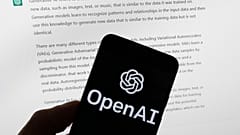 The OpenAI logo is displayed on a mobile phone in front of a computer screen with output from ChatGPT.