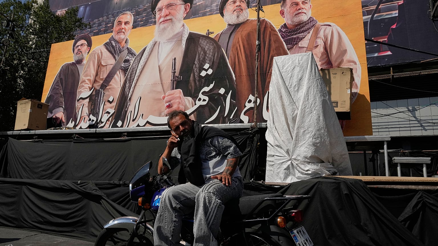 رجل يتحدث عبر الهاتف أمام لوحة تضم صور المرشد الإيراني السابق علي خامنئي وقادة إيرانيين ولبنانيين سابقين، في ساحة الثورة بالعاصمة طهران، 12 أكتوبر 2026. (أ ب)