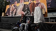 رجل يتحدث عبر الهاتف أمام لوحة تضم صور المرشد الإيراني السابق علي خامنئي وقادة إيرانيين ولبنانيين سابقين، في ساحة الثورة بالعاصمة طهران، 12 أكتوبر 2026. (أ ب)