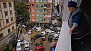 Moradores locais caminham entre veículos virados e destroços espalhados após as inundações provocadas pela chuva intensa.
