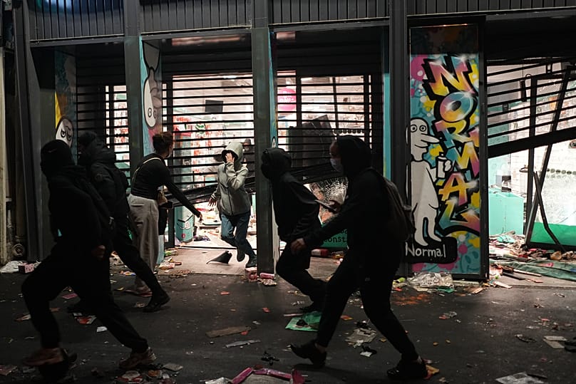 Protesters run past a looted store during a demonstration against rising living costs, joining students' grievances, in Paris, Saturday, Oct. 17, 2026.