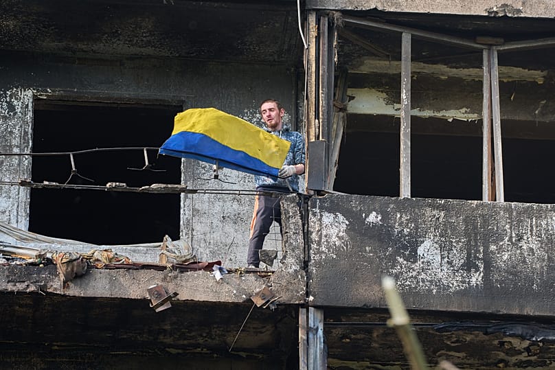 A man hangs out the national flag at his damaged apartment balcony after a missile strike during Russia's night air attack in Kyiv, 14 October, 2026