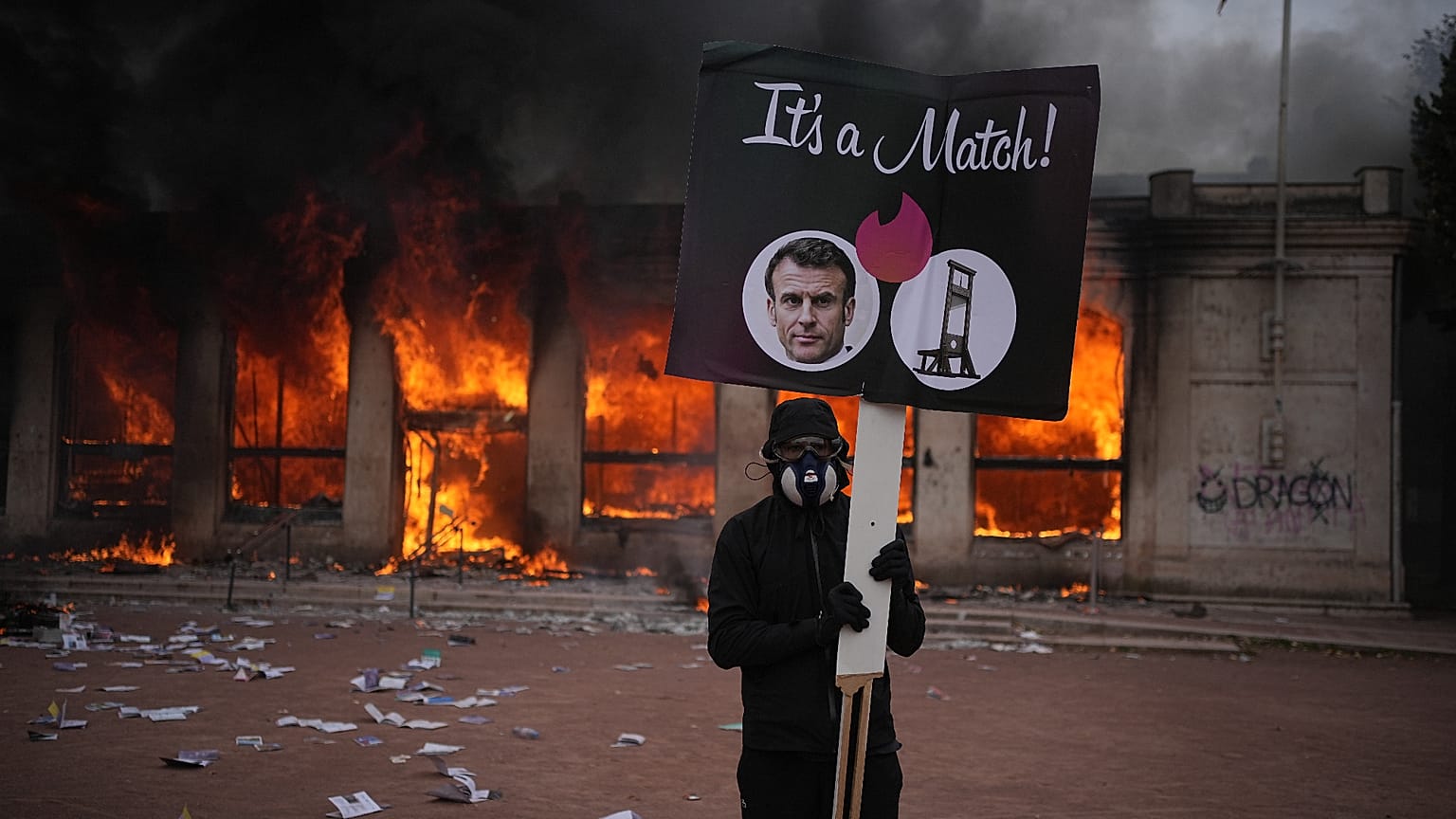 Fransa'nın Lyon kentinde 17 Ekim 2026 Cumartesi günü, geçim maliyeti protestosunda bir gösterici, yanan turizm ofisi önünde Emmanuel Macron fotoğraflı pankart taşıyor.