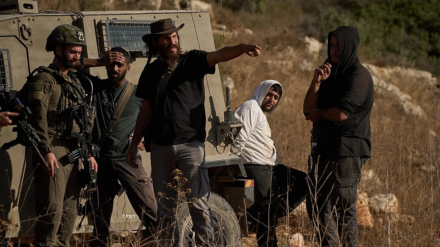 مستوطن إسرائيلي مسلح يتحدث إلى جندي، بينما يظهر رجل فلسطيني (ثاني شخص من اليمين) قيد الاحتجاز