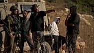 مستوطن إسرائيلي مسلح يتحدث إلى جندي، بينما يظهر رجل فلسطيني (ثاني شخص من اليمين) قيد الاحتجاز
