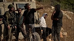 مستوطن إسرائيلي مسلح يتحدث إلى جندي، بينما يظهر رجل فلسطيني (ثاني شخص من اليمين) قيد الاحتجاز