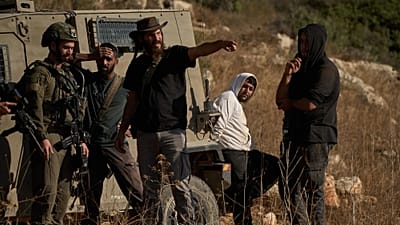 مستوطن إسرائيلي مسلح يتحدث إلى جندي، بينما يظهر رجل فلسطيني (ثاني شخص من اليمين) قيد الاحتجاز